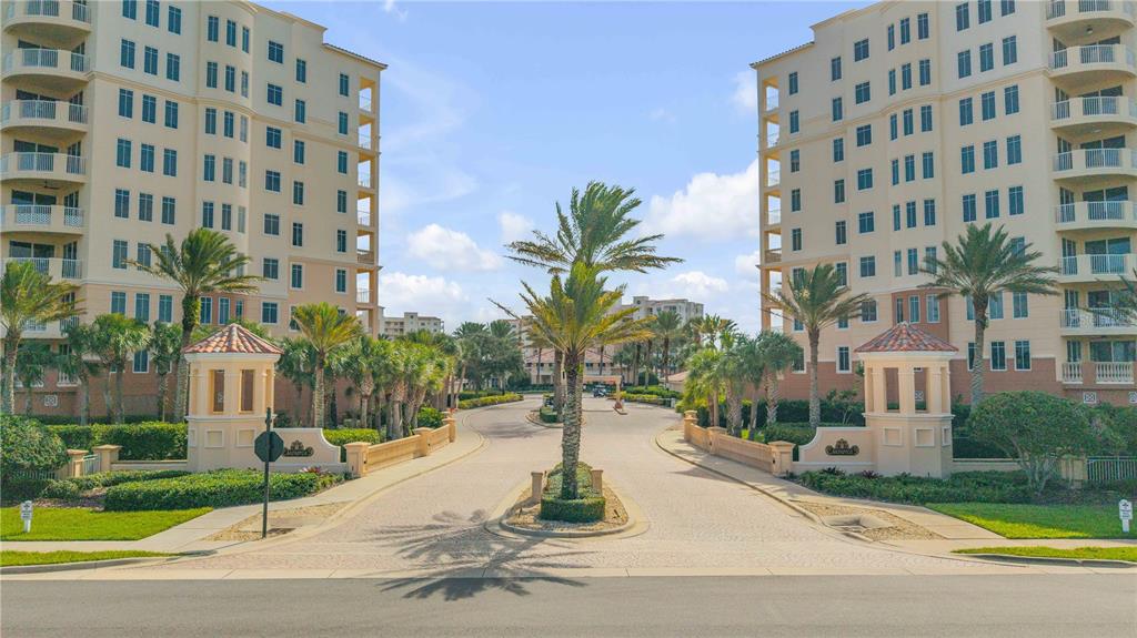 A gated entrance with two tall residential buildings, palm trees, and a paved driveway — 261 MINORCA BEACH WAY UNIT 406, NEW SMYRNA BEACH, FL