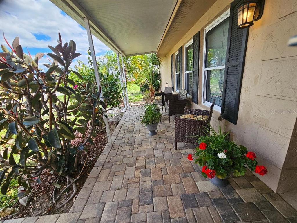 A paved patio features wicker chairs, potted plants, and a covered walkway along a house — 708 BRANNEN CIRCLE, SUN CITY CENTER, FL