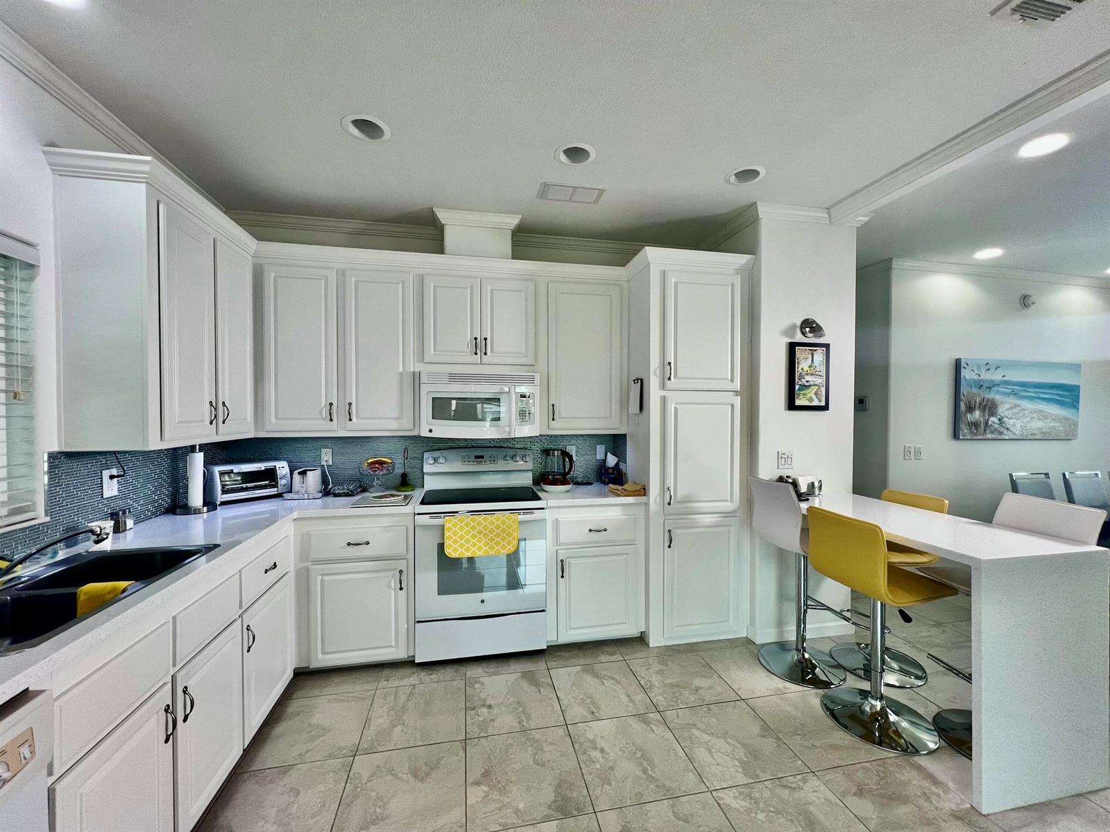 A white kitchen features a breakfast bar with yellow stools and light-colored tile flooring — 413 Hibiscus Drive, Deerfield Beach FL 33442, DEERFIELD BEACH, FL