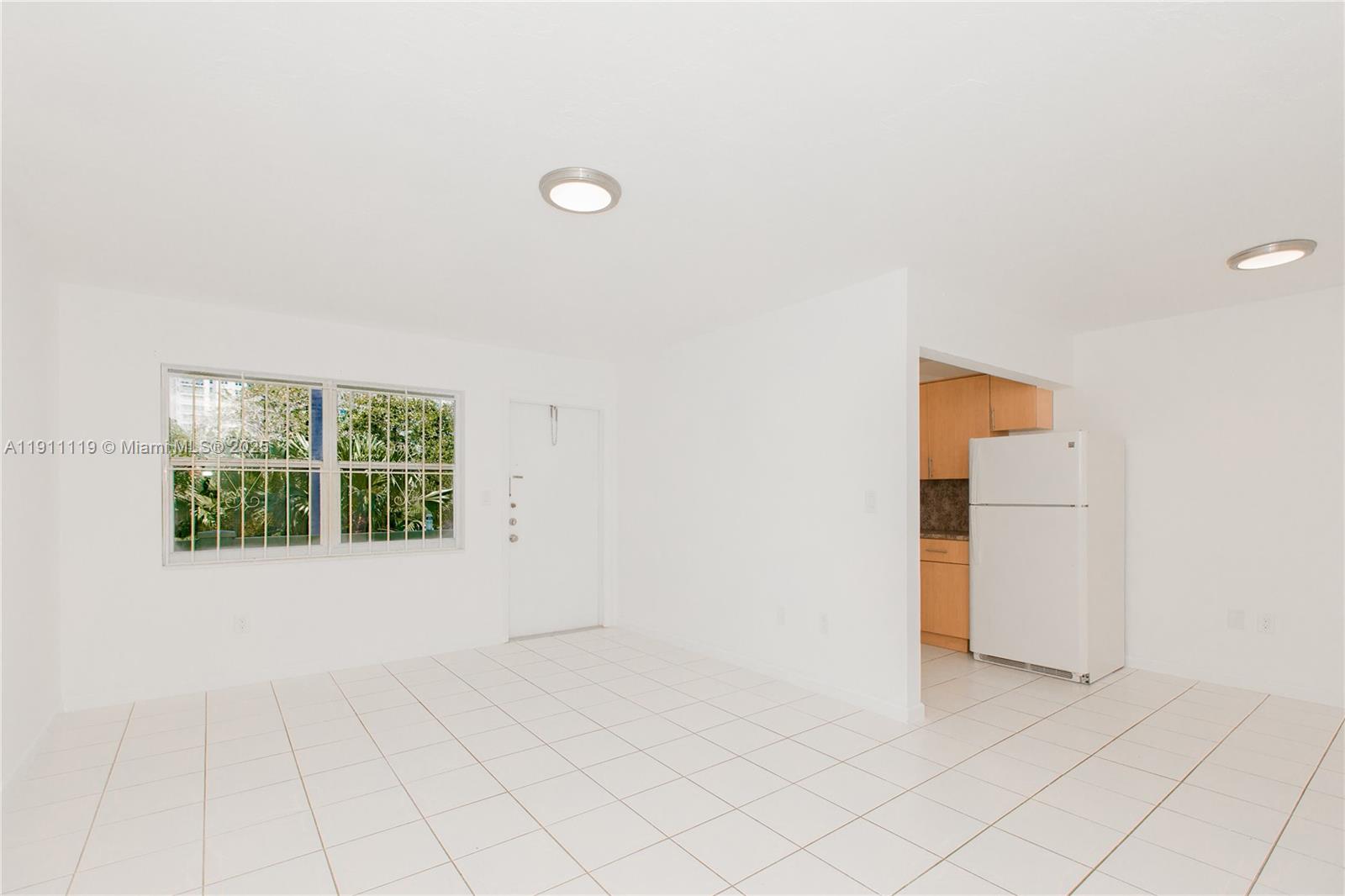 A bright, tiled living area features a white door, a window with security bars, and a kitchen — 1060 NE 78th St Rd # 10, Miami FL 33138, MIAMI, FL