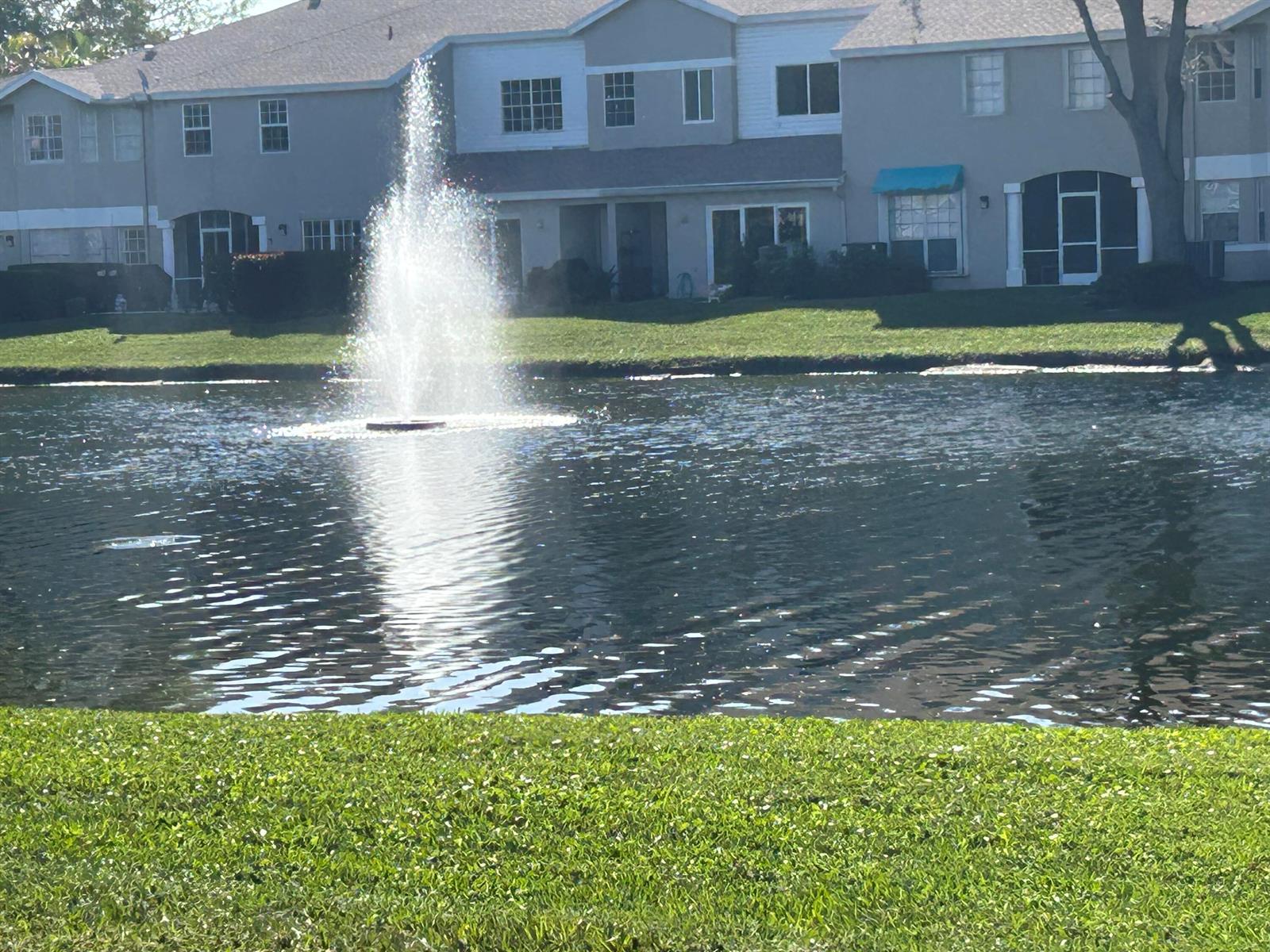 A pond with a fountain sits in front of multi-story apartment buildings under clear skies — 9082 Boca Gardens Parkway # C, Boca Raton FL 33496, BOCA RATON, FL