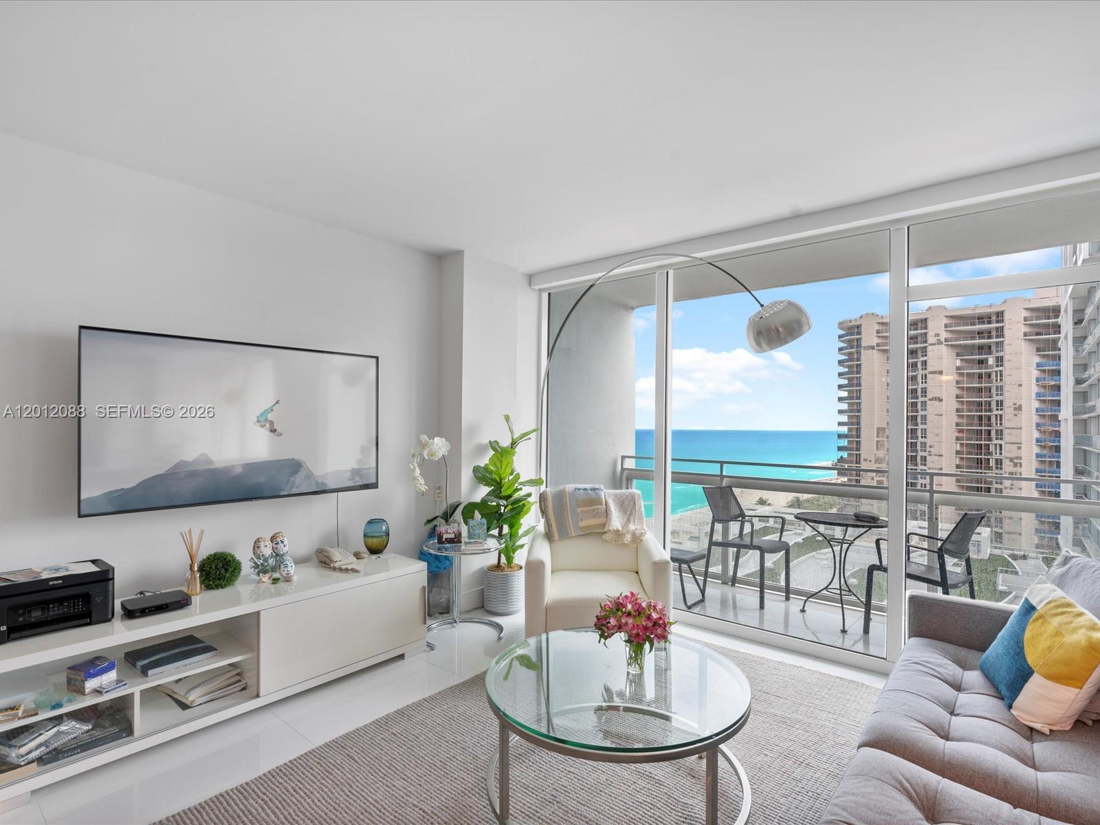 A modern living room features a glass coffee table, white media console, and balcony ocean views — 6801 Collins Ave # 1208, Miami Beach FL 33141, MIAMI BEACH, FL
