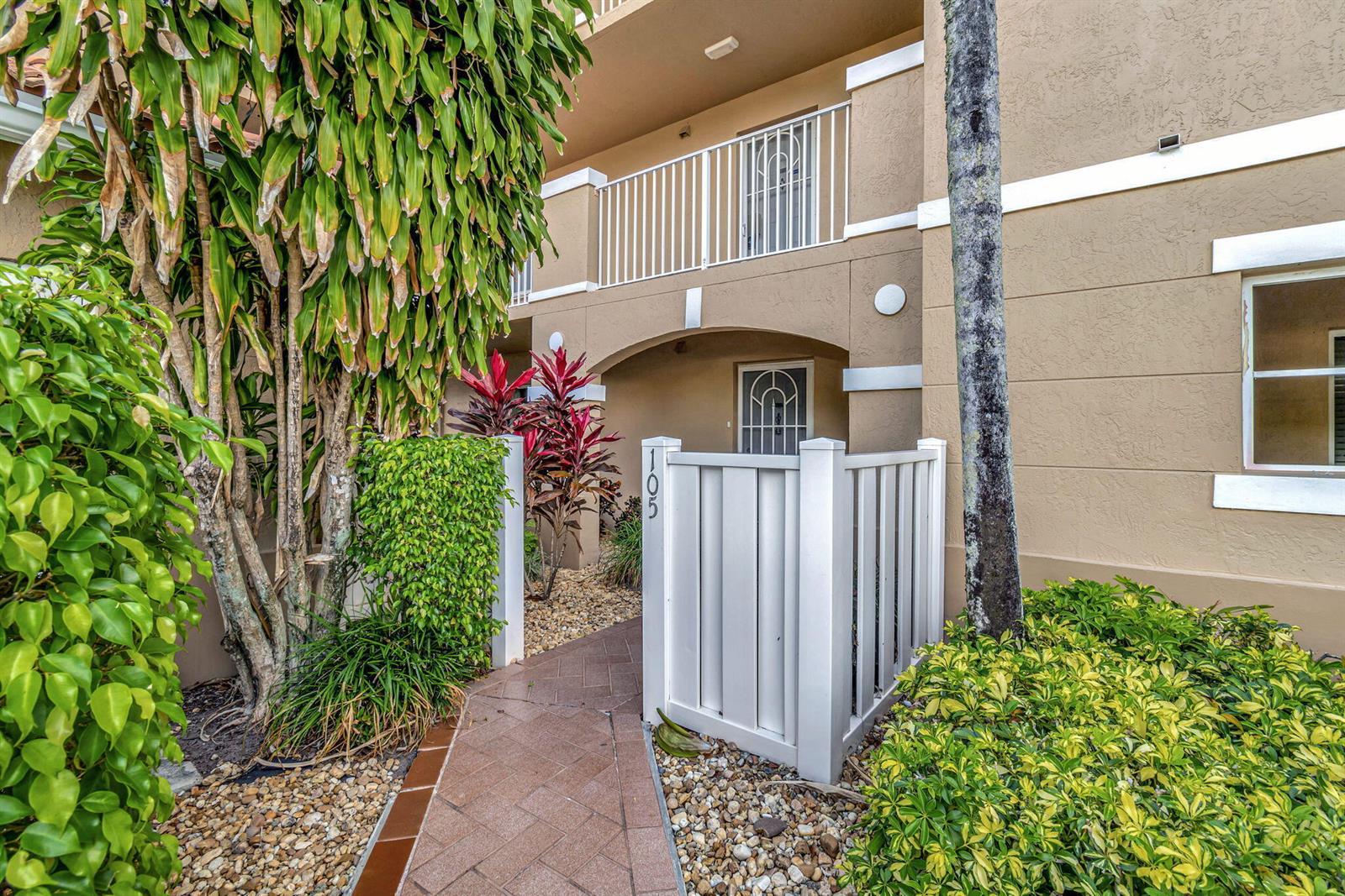 A paved walkway leads to the gated entrance of a tan building unit numbered 105 — 5842 Crystal Shores Drive # 105, Boynton Beach FL 33437, BOYNTON BEACH, FL