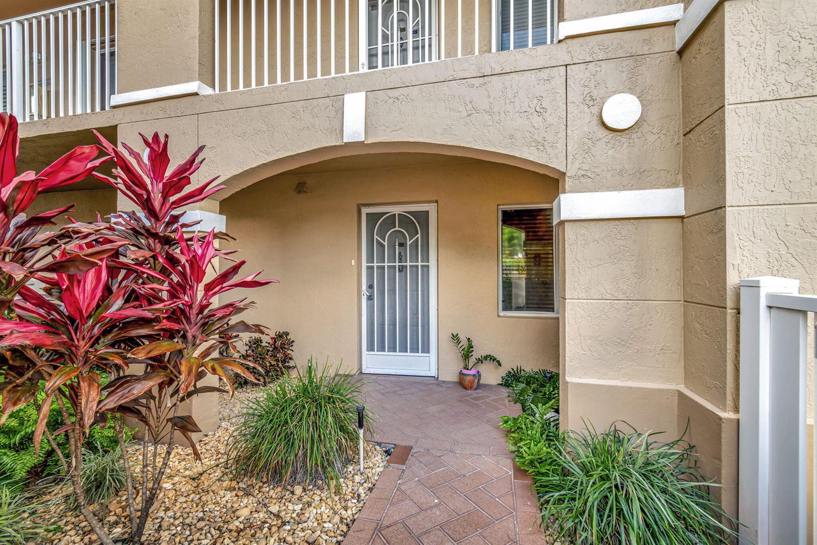 A tan building exterior features a covered entryway with a white storm door and walkway — 5842 Crystal Shores Drive # 105, Boynton Beach FL 33437, BOYNTON BEACH, FL