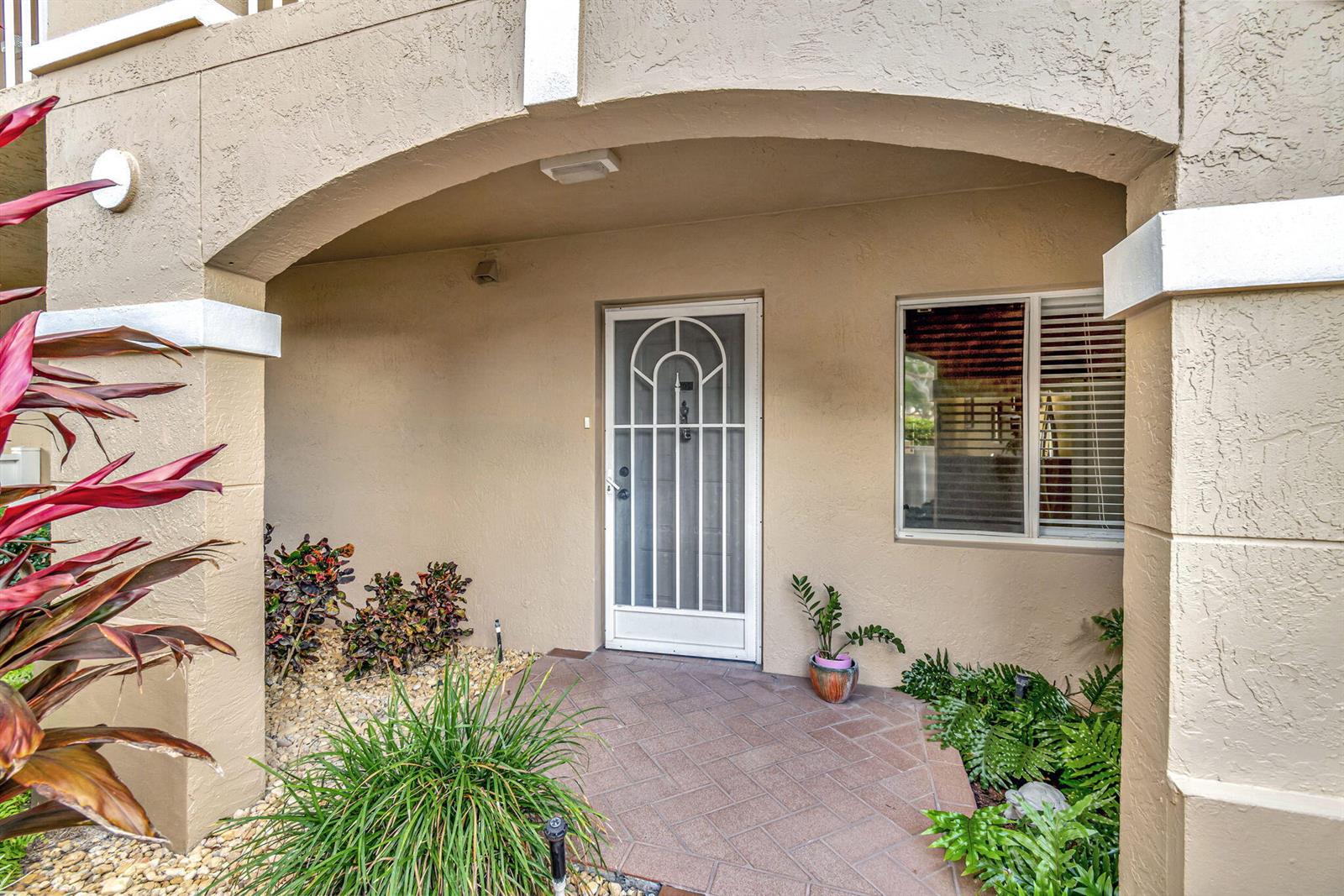 A covered entryway features a white storm door, a window with blinds, and landscaped plants — 5842 Crystal Shores Drive # 105, Boynton Beach FL 33437, BOYNTON BEACH, FL