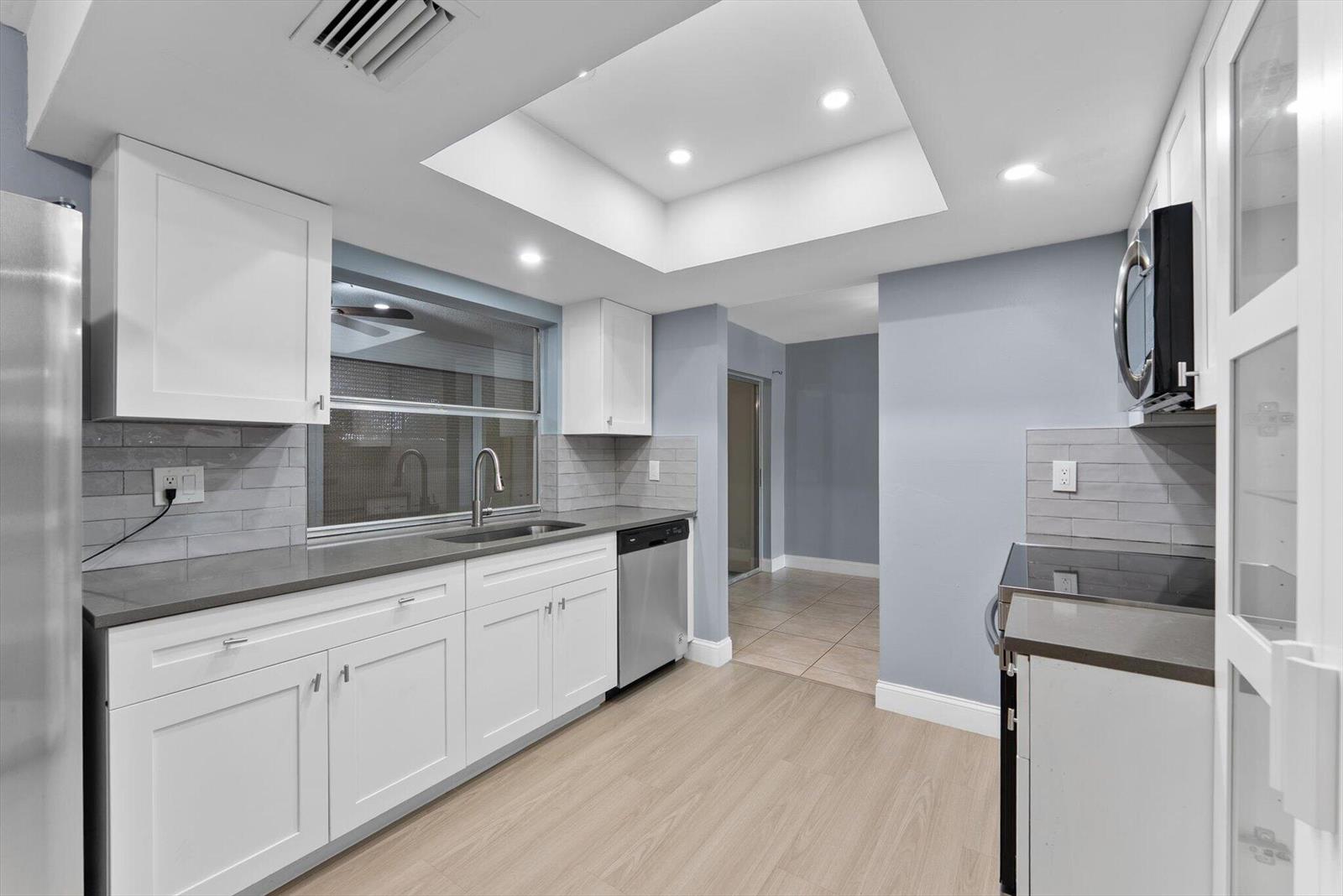 A modern kitchen features white cabinets, gray countertops, stainless steel appliances, and light wood flooring — 901 NW 49th Street, Deerfield Beach FL 33064, DEERFIELD BEACH, FL
