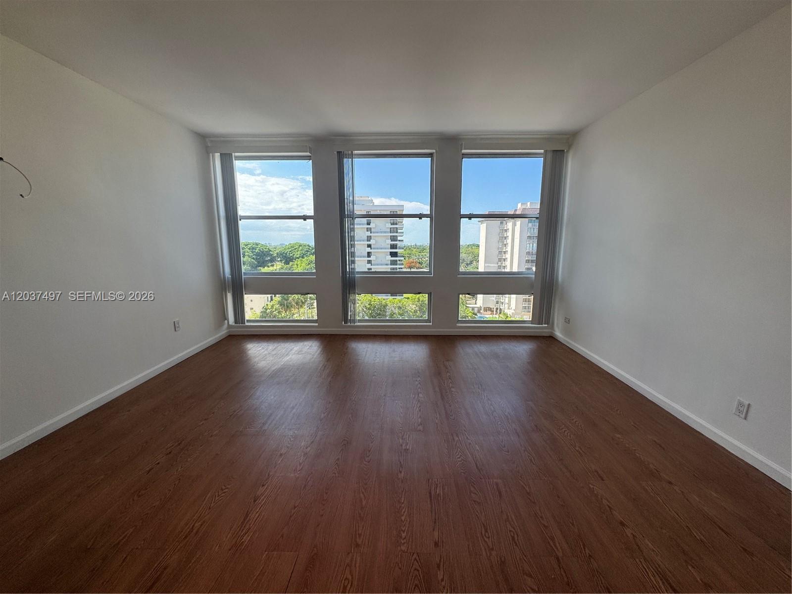 An empty room features wood-look flooring, white walls, and three large windows overlooking buildings — 700 Biltmore Way # 916, Coral Gables FL 33134, CORAL GABLES, FL
