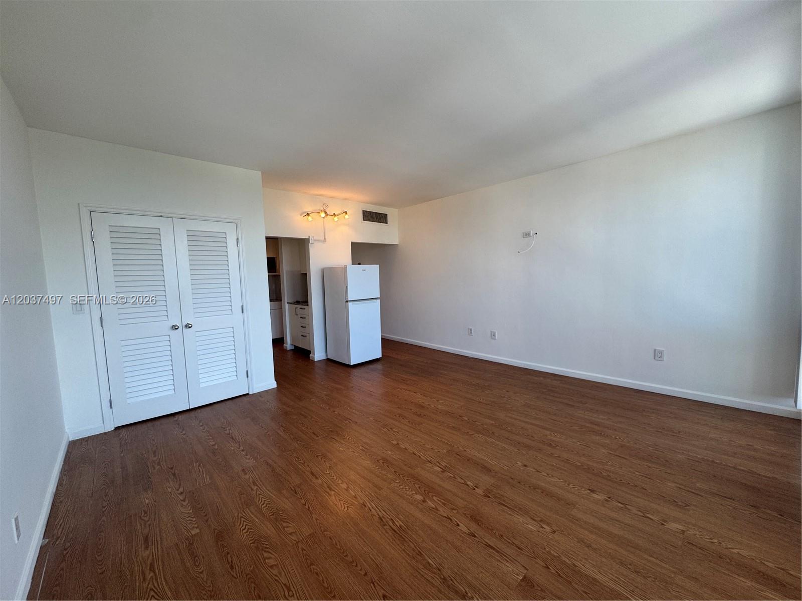 An empty room features wood-look flooring, white walls, a white refrigerator, and louvered closet doors — 700 Biltmore Way # 916, Coral Gables FL 33134, CORAL GABLES, FL