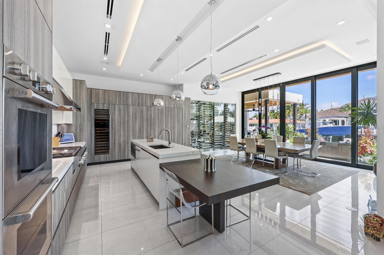A modern kitchen and dining area features wood cabinetry, a white island, and floor-to-ceiling windows — 2863 NE 27th Street, Fort Lauderdale FL 33306, FORT LAUDERDALE, FL
