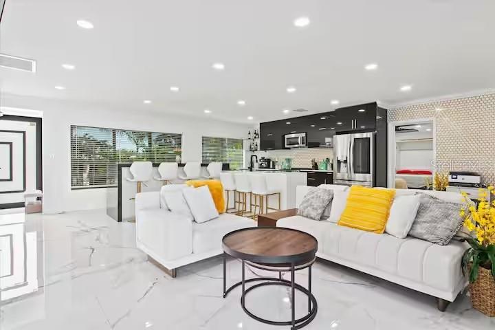 A modern open-concept living room and kitchen feature white marble floors and yellow accent pillows — 6612 9TH AVENUE NW, BRADENTON, FL