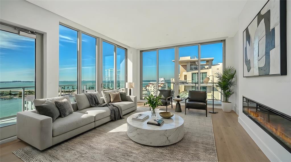 A modern living room features a marble coffee table, fireplace, and floor-to-ceiling windows with water views — 253 GOLDEN GATE POINT UNIT 902, SARASOTA, FL