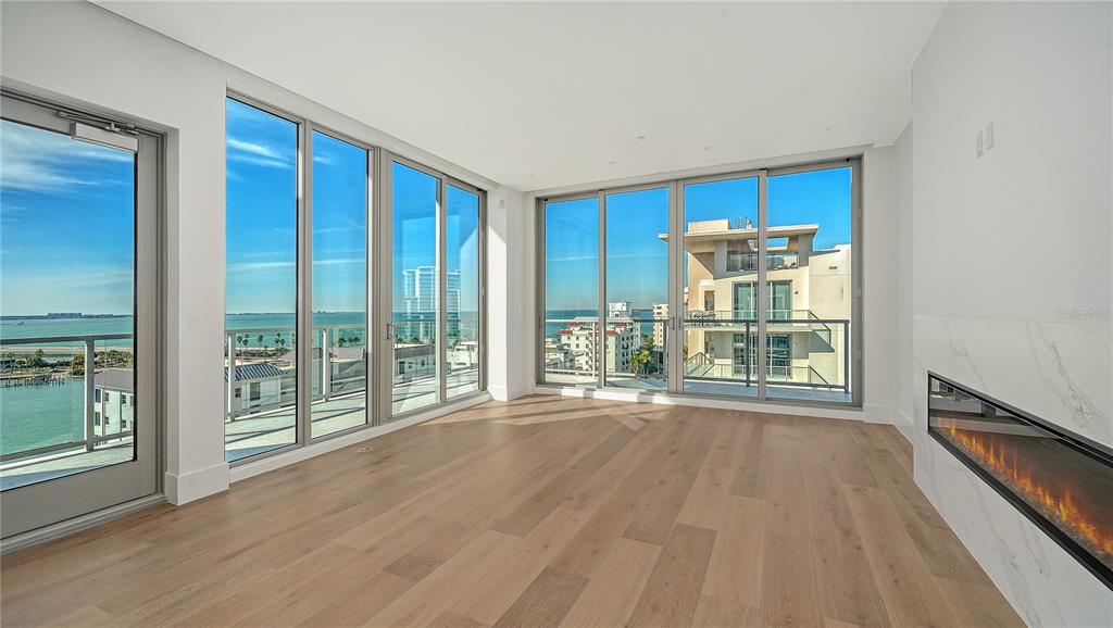 A modern living room with light wood floors, a fireplace, and floor-to-ceiling windows overlooking water — 253 GOLDEN GATE POINT UNIT 902, SARASOTA, FL