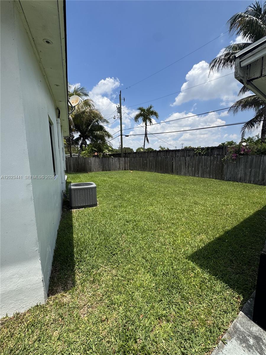 A grassy backyard features a white exterior wall, an air conditioning unit, and wooden fencing — 5701 SW 117th Ave, Miami FL 33183, MIAMI, FL