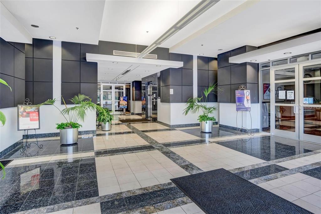 A modern building lobby features tiled floors, dark accent walls, and potted indoor plants — 6200 METROWEST BOULEVARD UNIT 103D, ORLANDO, FL
