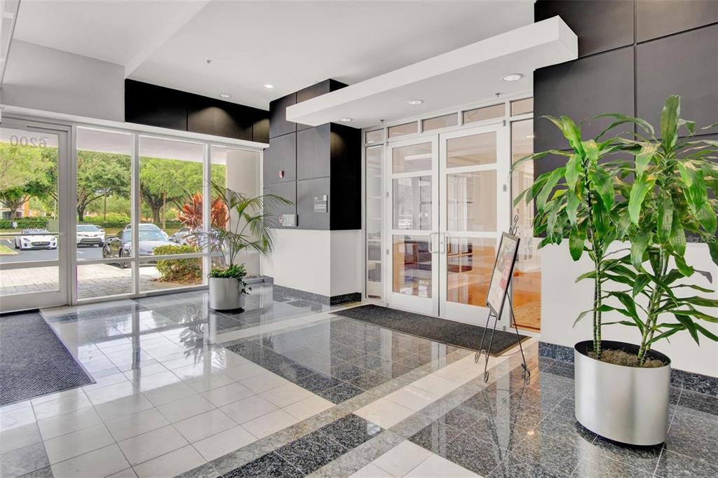 A modern building lobby features polished tile floors, glass entry doors, and two potted plants — 6200 METROWEST BOULEVARD UNIT 103D, ORLANDO, FL