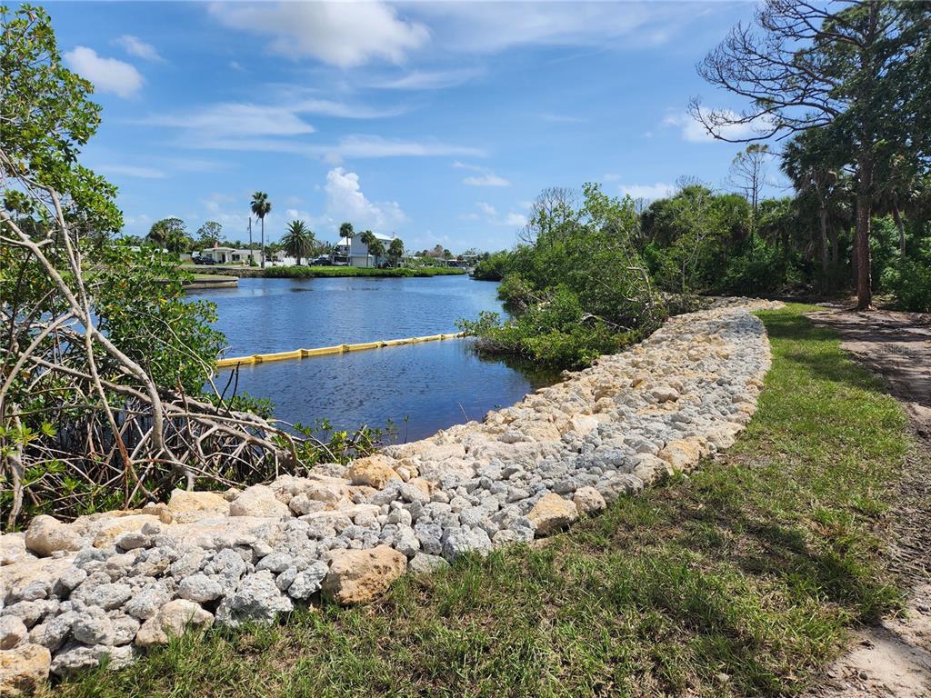 A rocky shoreline borders a calm canal with a yellow floating barrier under a blue sky — 971 LAMPP DRIVE, ENGLEWOOD, FL