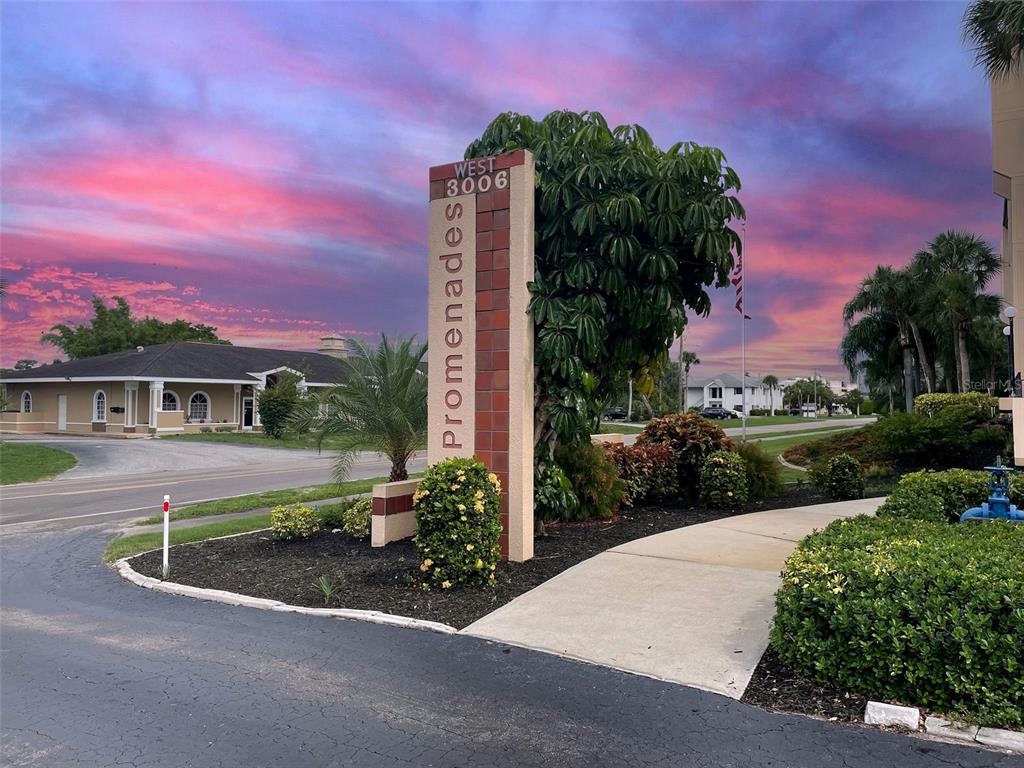 A tall, rectangular monument sign labeled Promenades stands near a paved road and sidewalk — 3006 CARING WAY UNIT 210, PORT CHARLOTTE, FL