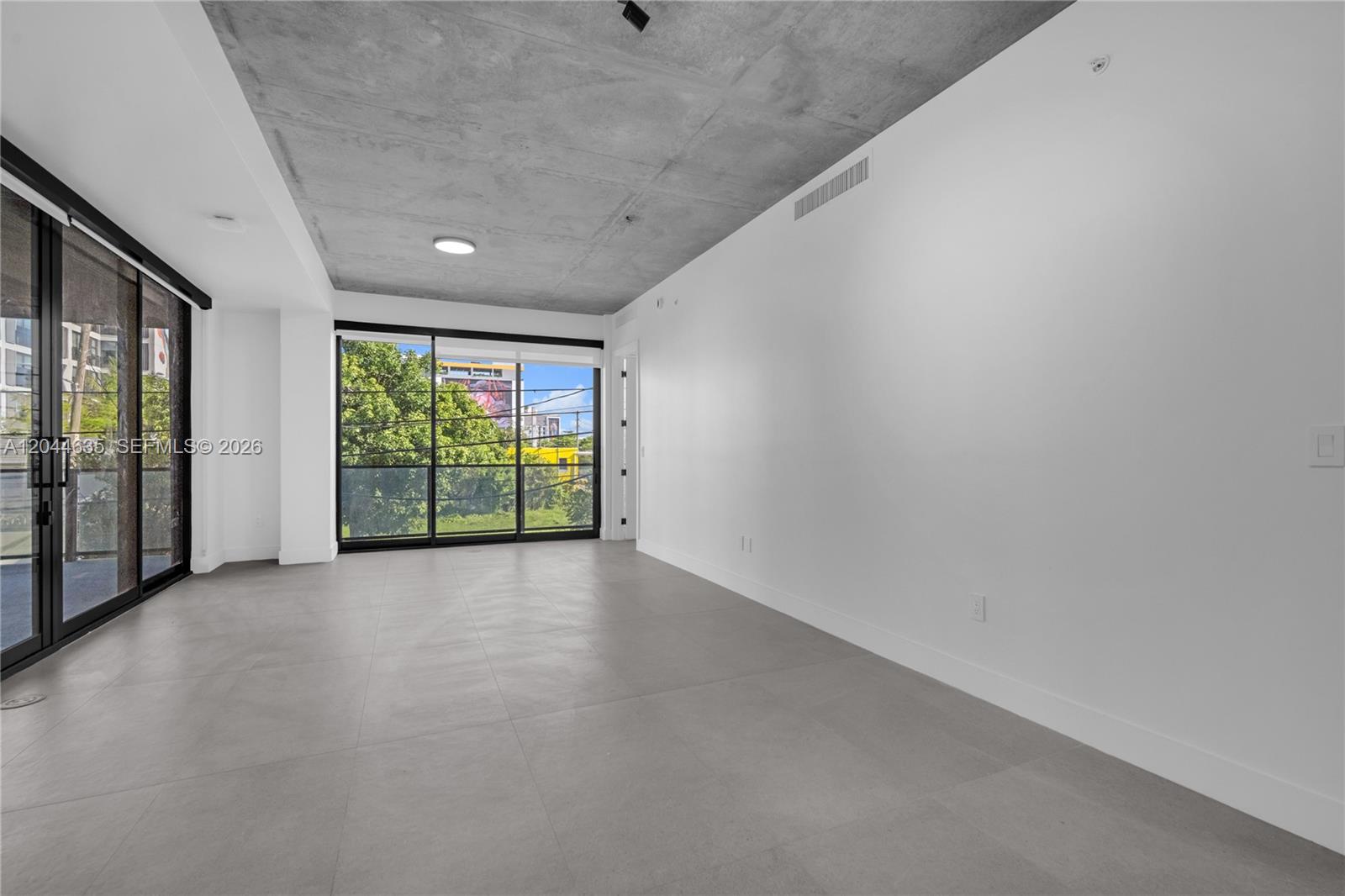 An unfurnished room features gray tile flooring, a concrete ceiling, and large sliding glass doors — 91 NW 27th St # 101, Miami FL 33127, MIAMI, FL
