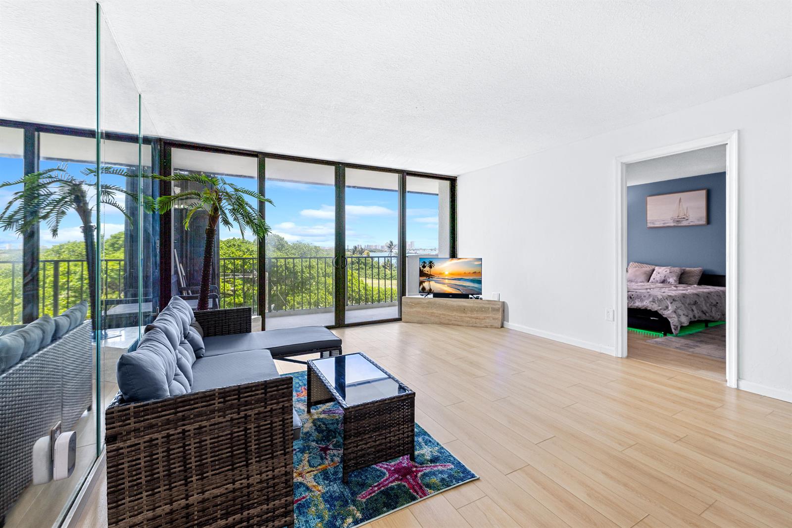A living room features light wood-look flooring, a wicker sectional, and floor-to-ceiling windows with balcony access — 5600 N Flagler Drive # 407, West Palm Beach FL 33407, WEST PALM BEACH, FL