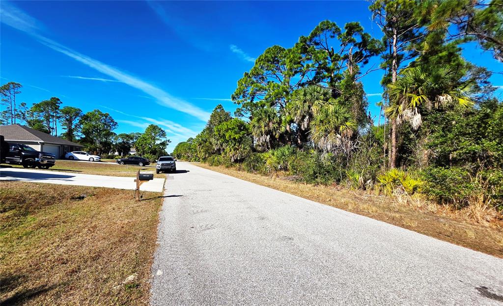 A paved street runs past a residential driveway and a wooded lot under a blue sky — 17492 WACO AVENUE, PORT CHARLOTTE, FL