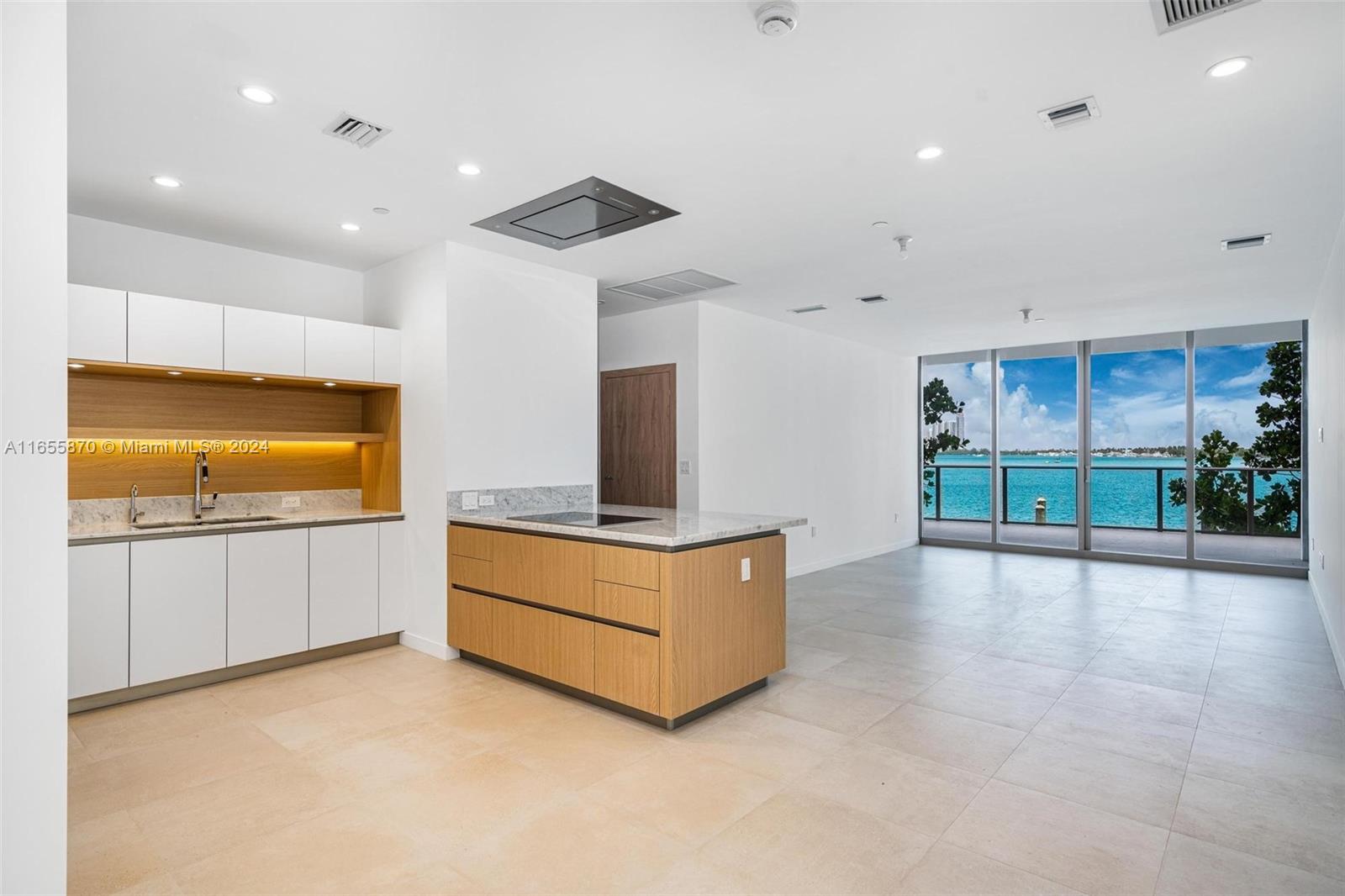 A modern kitchen with wood cabinetry and a marble island overlooks a waterfront balcony — 1135 103 St # 202, Bay Harbor Islands FL 33154, BAY HARBOR ISLANDS, FL