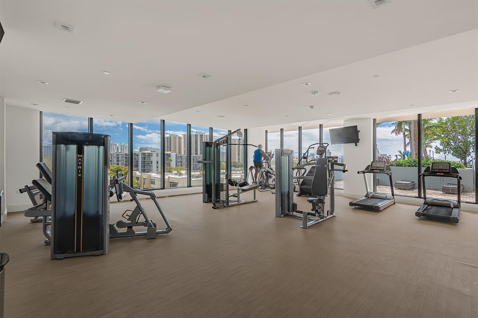 A gym with various exercise machines, light flooring, and floor-to-ceiling windows overlooking city buildings — 2960 NE 207th Street # 708, Aventura FL 33180, AVENTURA, FL