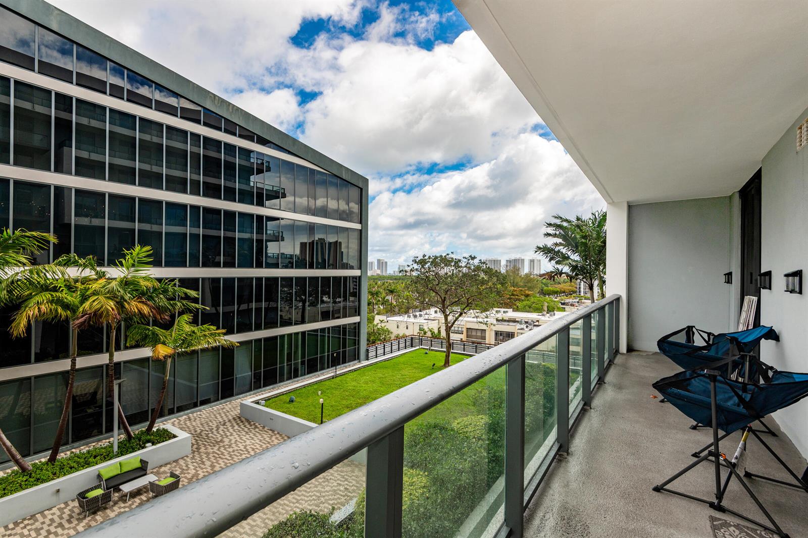 A balcony with two folding chairs overlooks a green courtyard and a modern glass building — 2960 NE 207th Street # 708, Aventura FL 33180, AVENTURA, FL