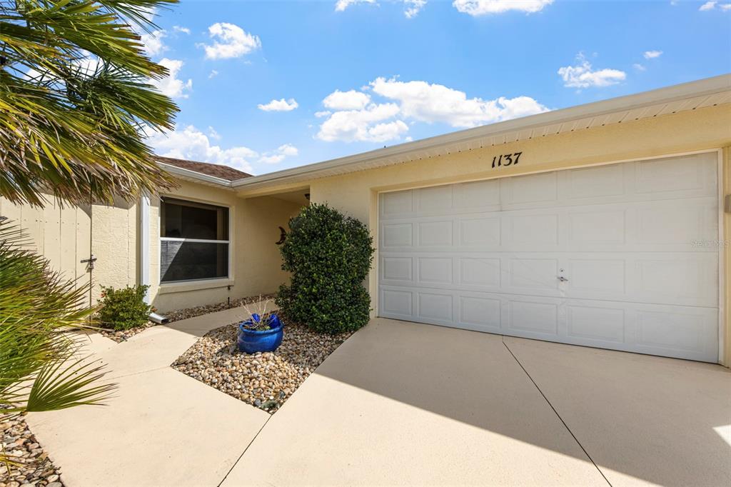 A beige house exterior features a white garage door, a paved driveway, and a small garden — 1137 SANTA CRUZ DRIVE, THE VILLAGES, FL