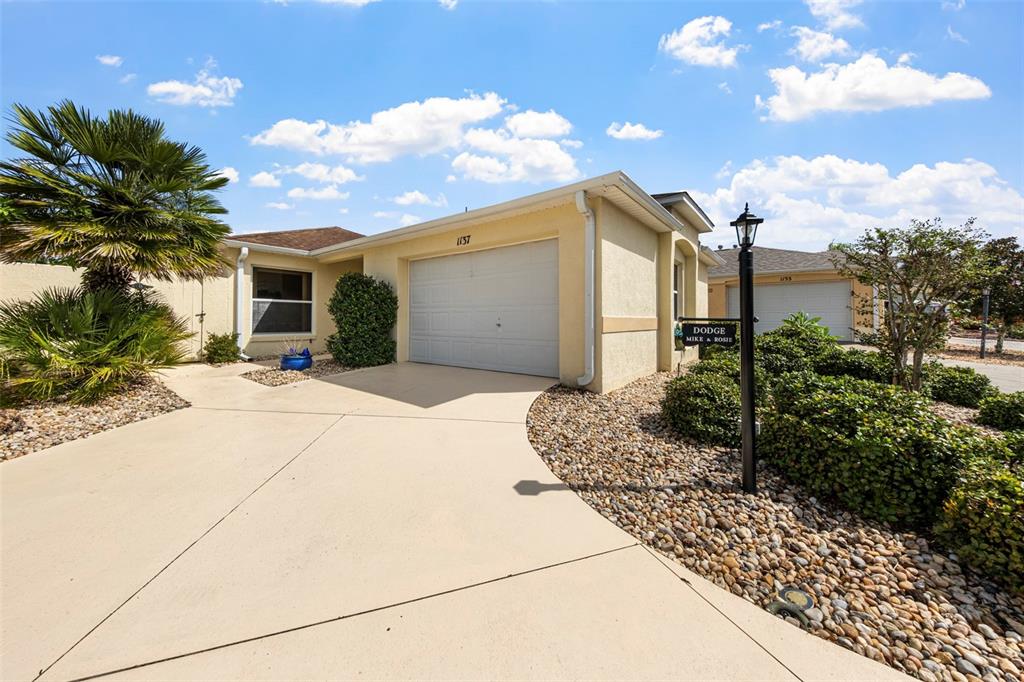 A beige single-story house with a two-car garage, a palm tree, and a rock garden — 1137 SANTA CRUZ DRIVE, THE VILLAGES, FL