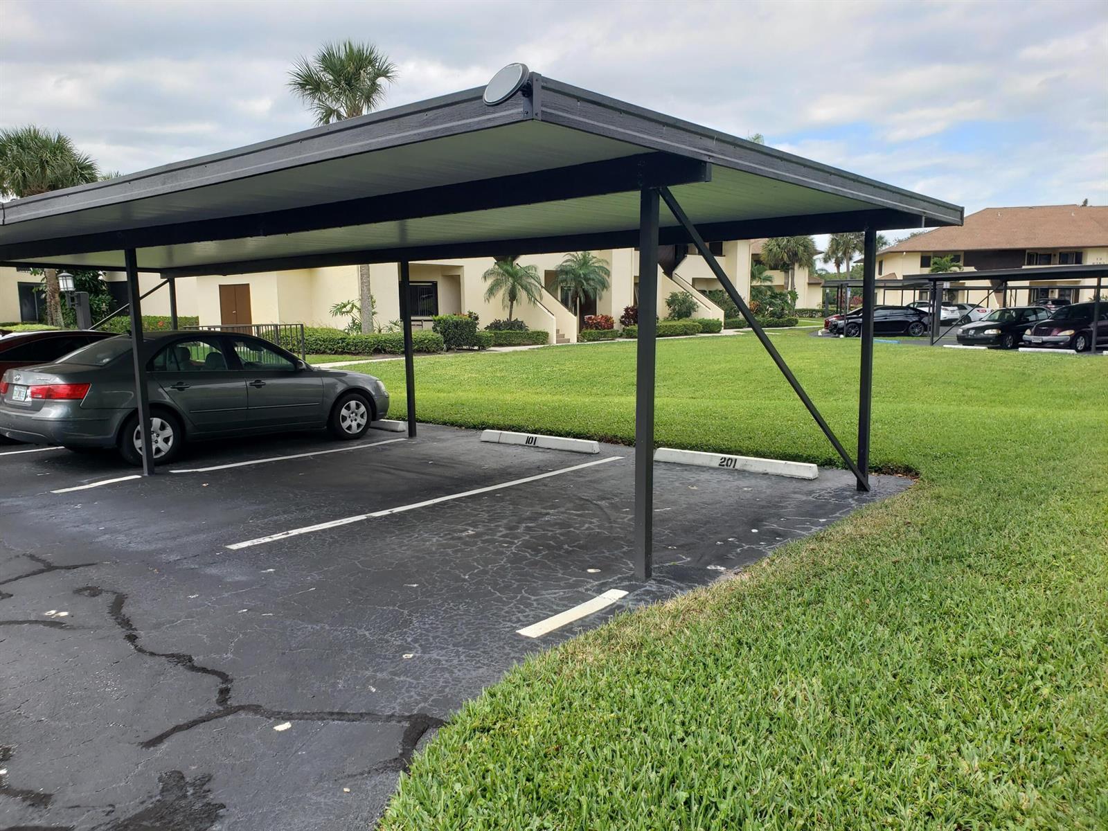 A covered parking carport with numbered spaces sits in front of a multi-story apartment building — 6303 SE Williamsburg Drive # 201, Hobe Sound FL 33455, HOBE SOUND, FL