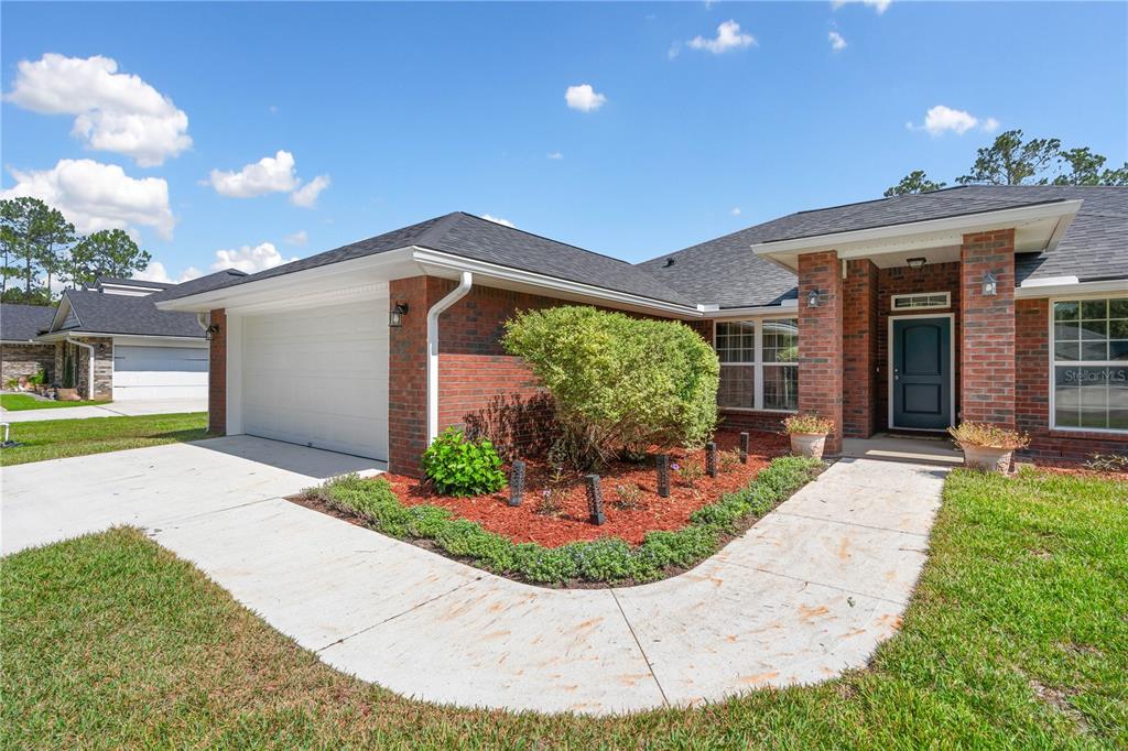 A brick-front house with a white garage door, a front walkway, and a landscaped garden — 4415 TALL PINES LANE, MIDDLEBURG, FL