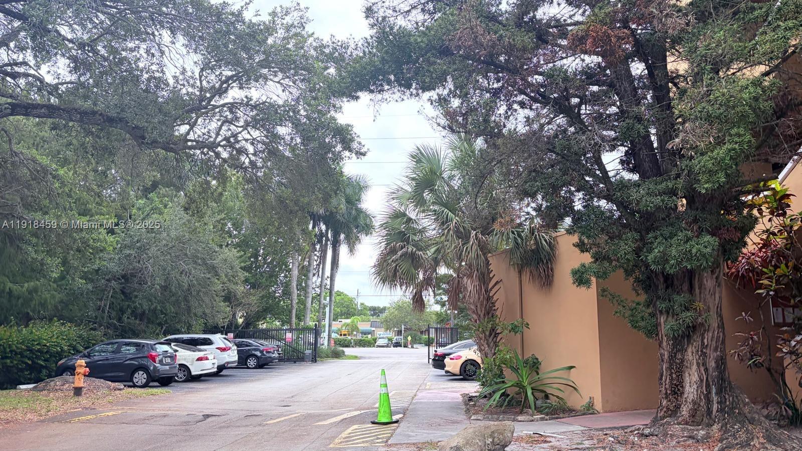 An outdoor parking area features several parked cars, mature trees, and a tan building wall — 4111 Stirling Rd # 102, Dania Beach FL 33314, DANIA BEACH, FL