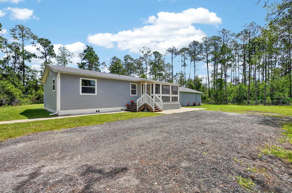 A gray single-story house with a screened porch and gravel driveway sits near a forest — 3798 BREEZY COURT, ORMOND BEACH, FL