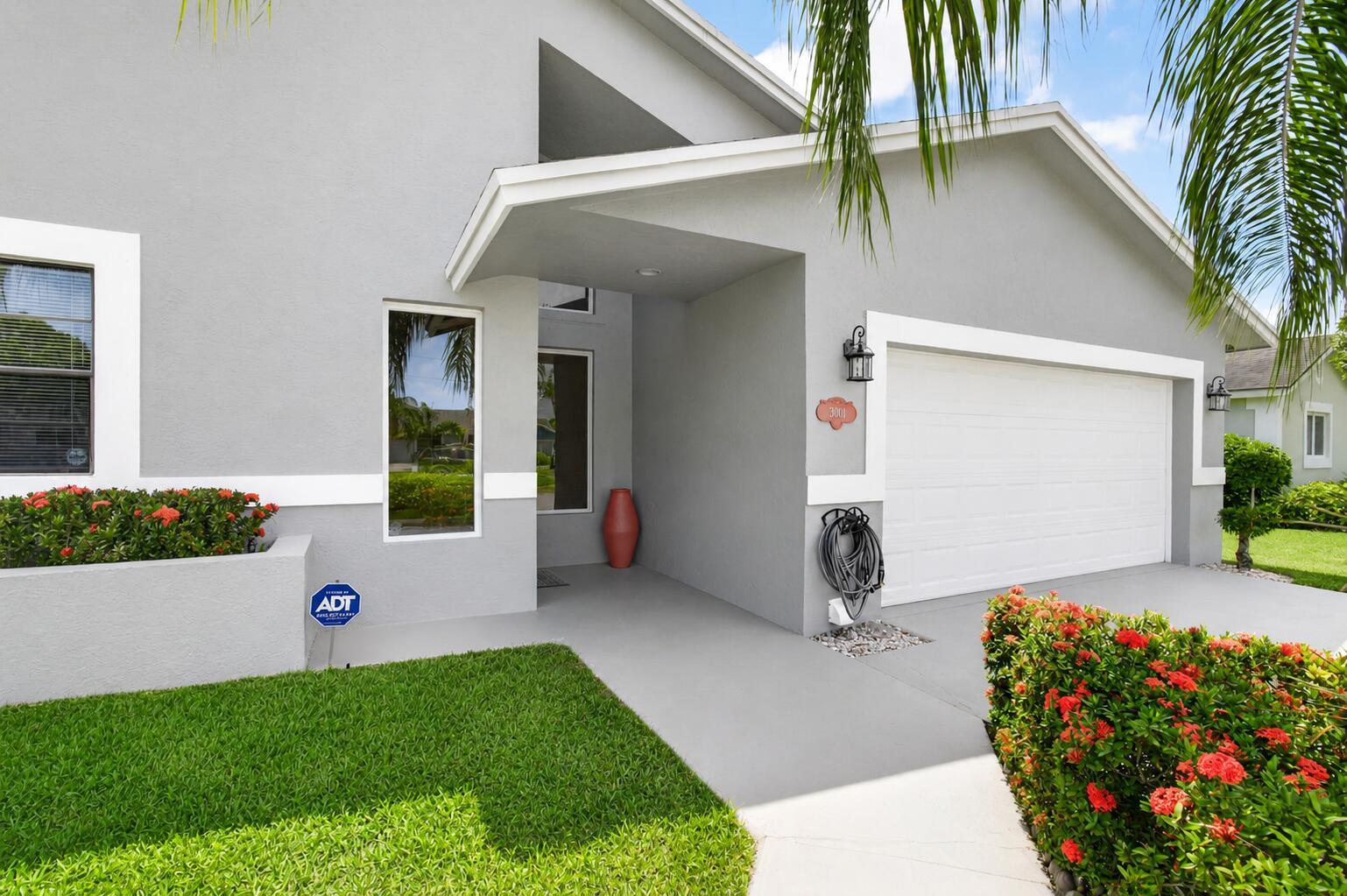 A gray house exterior features a white garage door, a covered entryway, and red flowers — 3001 Eagle Drive, Delray Beach FL 33444, DELRAY BEACH, FL