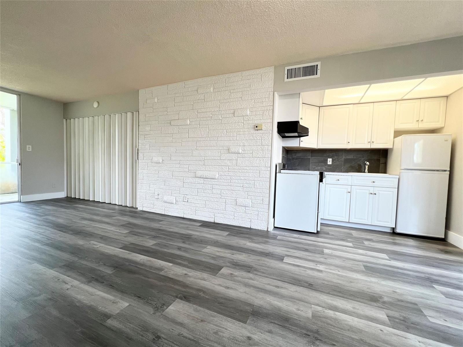 A room with gray wood-look flooring, a white brick accent wall, and a small kitchen — 777 SE 2nd Avenue # A102, Deerfield Beach FL 33441, DEERFIELD BEACH, FL