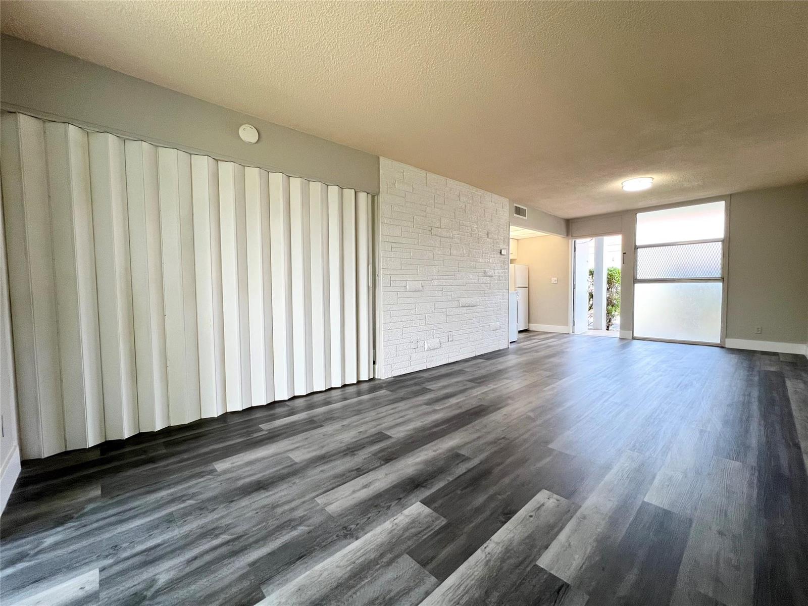 An empty room features gray wood-look flooring, a white brick accent wall, and folding doors — 777 SE 2nd Avenue # A102, Deerfield Beach FL 33441, DEERFIELD BEACH, FL