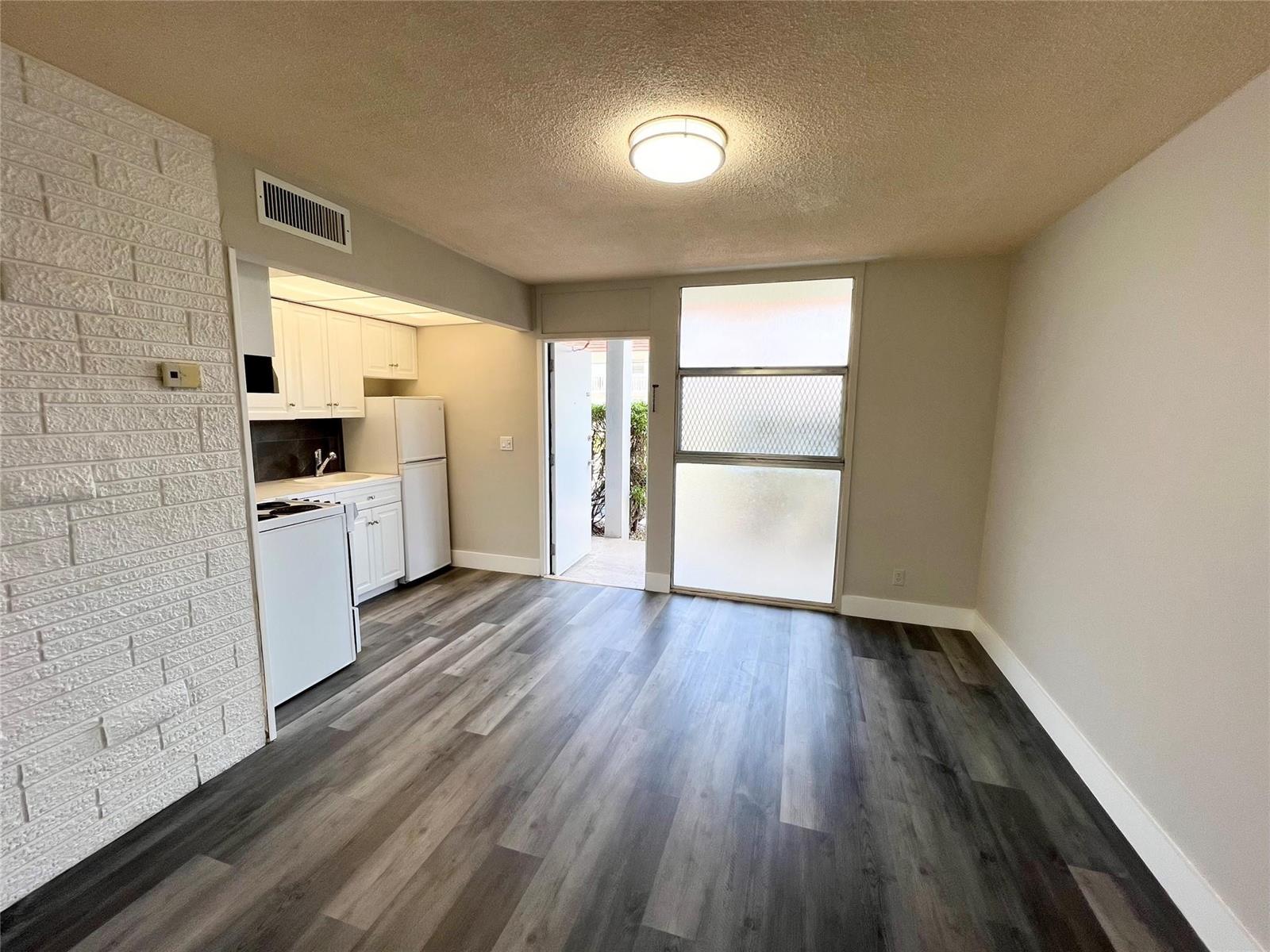 A room with wood-look flooring, a white brick wall, and an adjacent small kitchen area — 777 SE 2nd Avenue # A102, Deerfield Beach FL 33441, DEERFIELD BEACH, FL