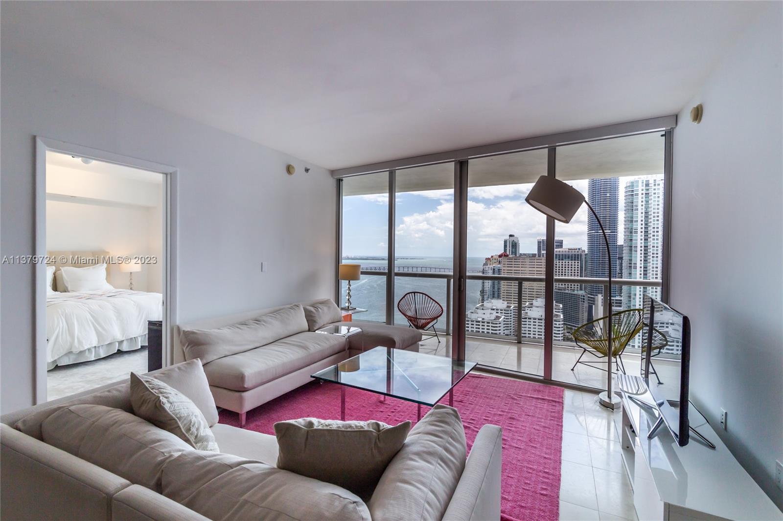 A living room with a pink rug, glass coffee table, and balcony overlooking a city — 495 Brickell Ave # 3603, Miami FL 33131, MIAMI, FL