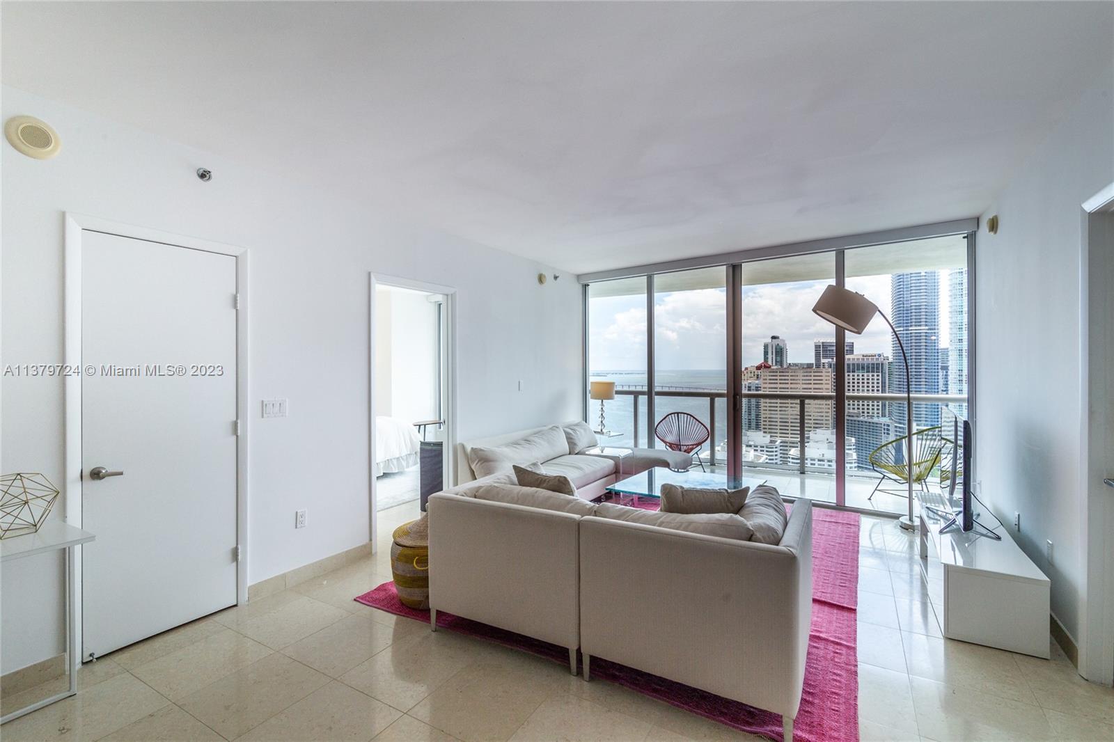 A living room features a sectional sofa, a pink rug, and floor-to-ceiling windows overlooking a city — 495 Brickell Ave # 3603, Miami FL 33131, MIAMI, FL