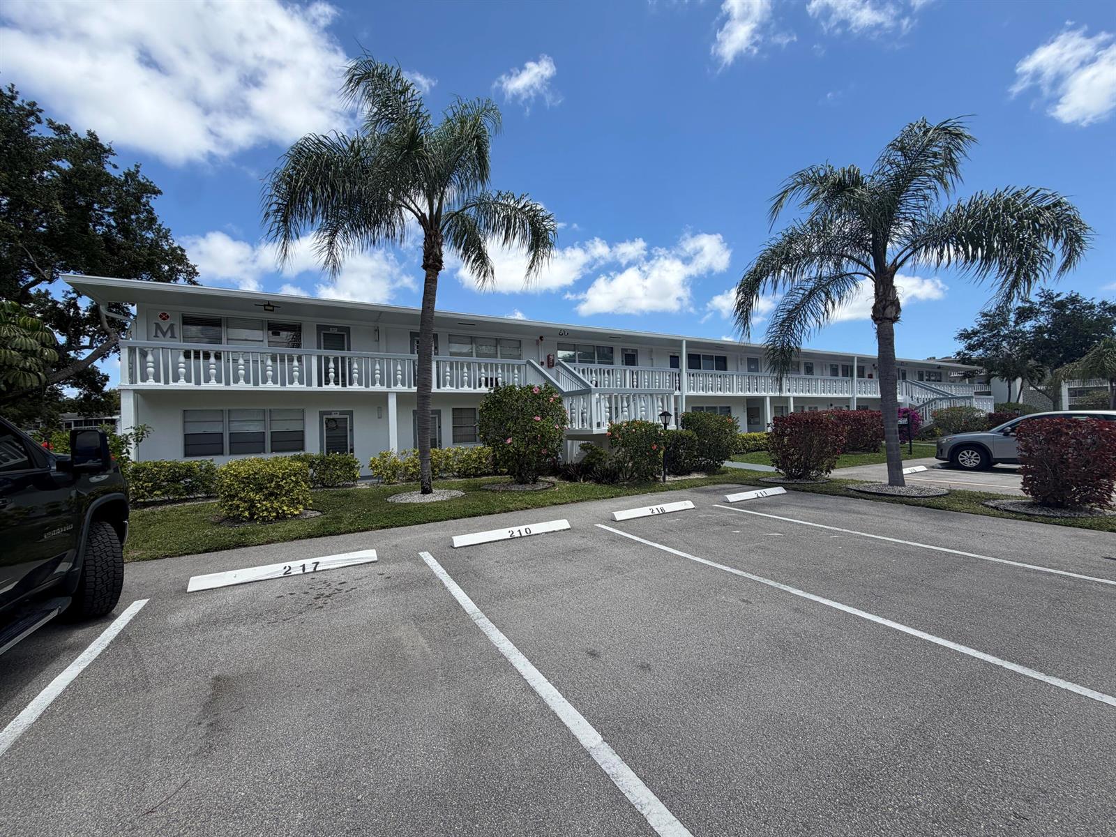 A two-story white apartment building features a balcony, palm trees, and numbered parking spaces — 209 Newport M # 209, Deerfield Beach FL 33442, DEERFIELD BEACH, FL