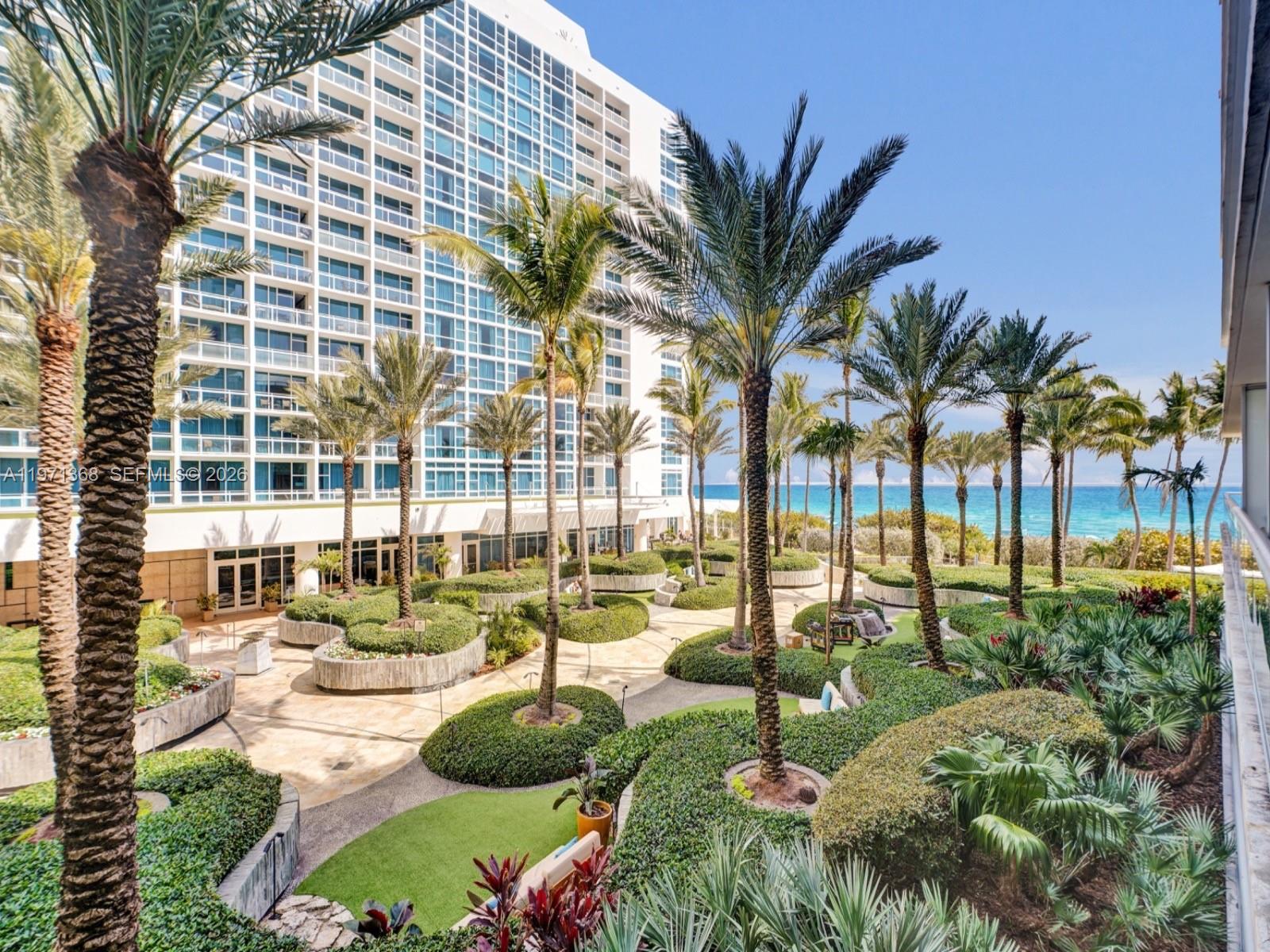 An elevated view of a landscaped courtyard with palm trees and a tall apartment building — 6799 Collins Ave # 201, Miami Beach FL 33141, MIAMI BEACH, FL