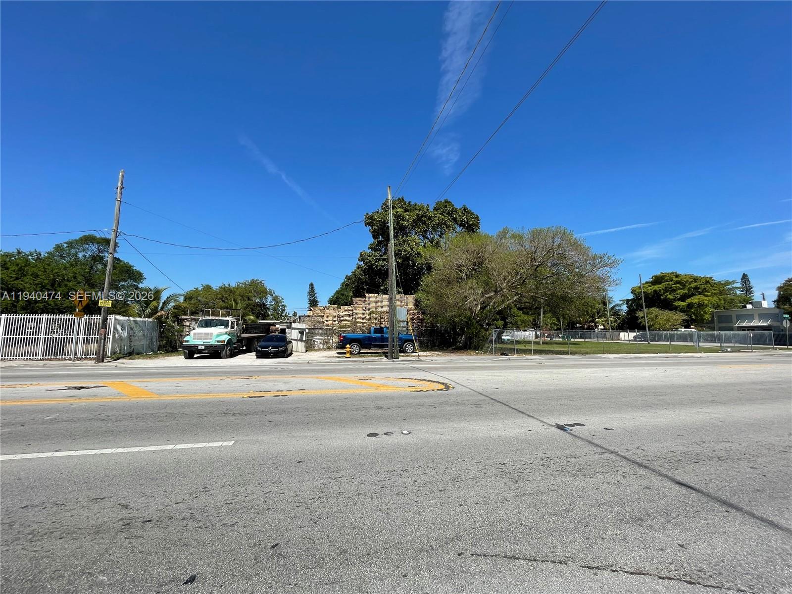 A paved road runs in front of a fenced lot containing stacked lumber and vehicles — 3021 NW 54th St, Miami FL 33142, MIAMI, FL