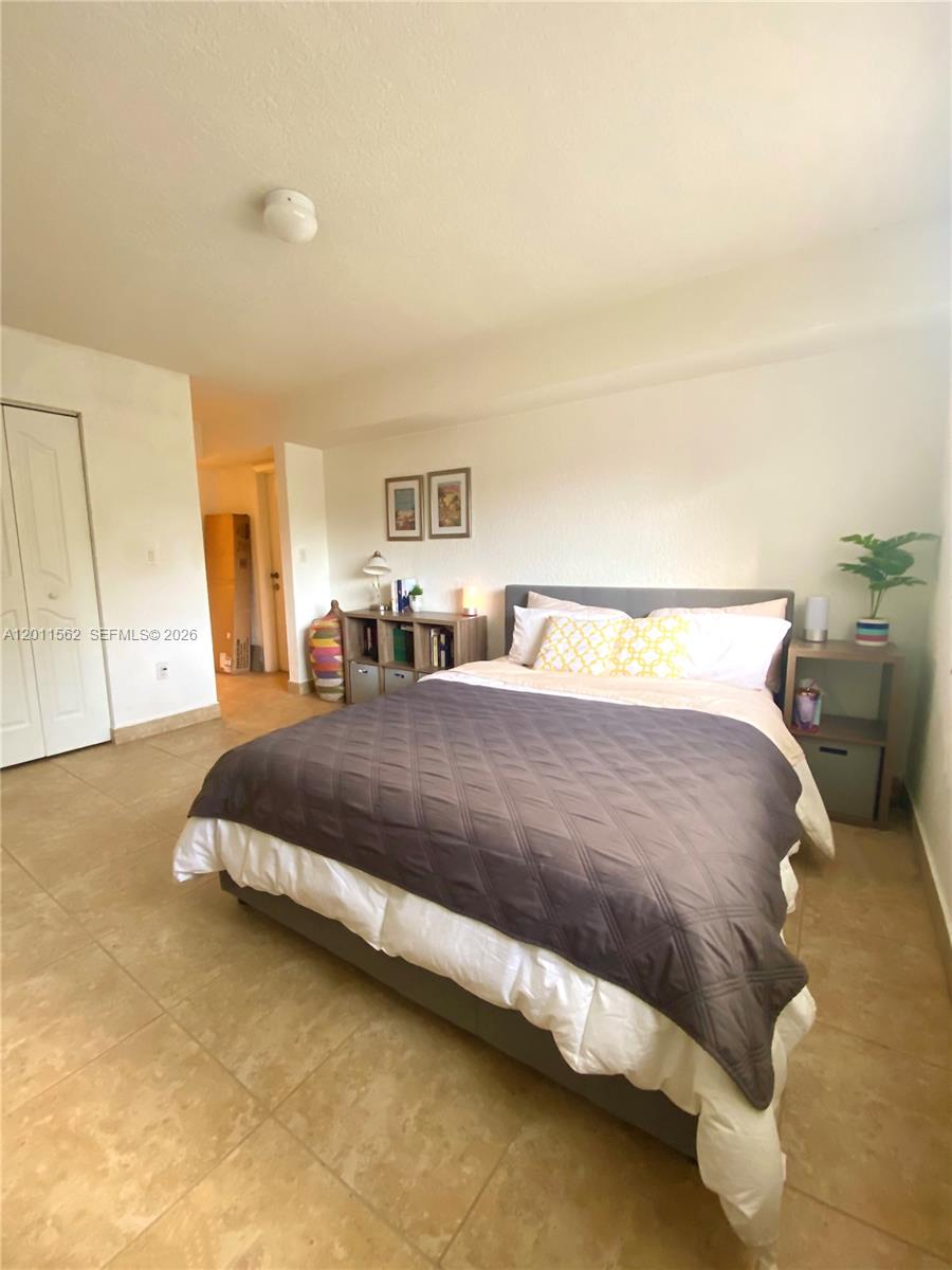 A bedroom features a bed with a dark quilt, tiled floors, and a small nightstand — 445 SW 11th St # 107, Miami FL 33130, MIAMI, FL