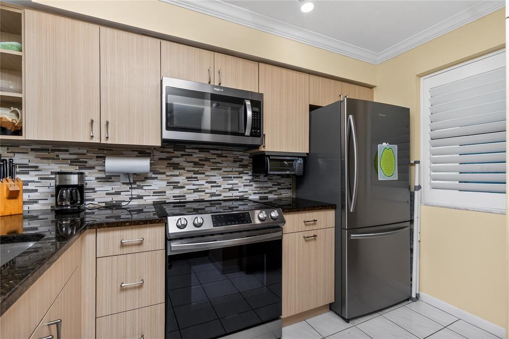 A kitchen features light wood cabinets, stainless steel appliances, a mosaic backsplash, and dark countertops — 1591 BEACH ROAD UNIT 407, ENGLEWOOD, FL