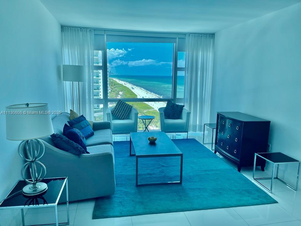 A living room with blue furniture and a rug overlooks the ocean through large windows — 6801 Collins Ave # C1115, Miami Beach FL 33141, MIAMI BEACH, FL