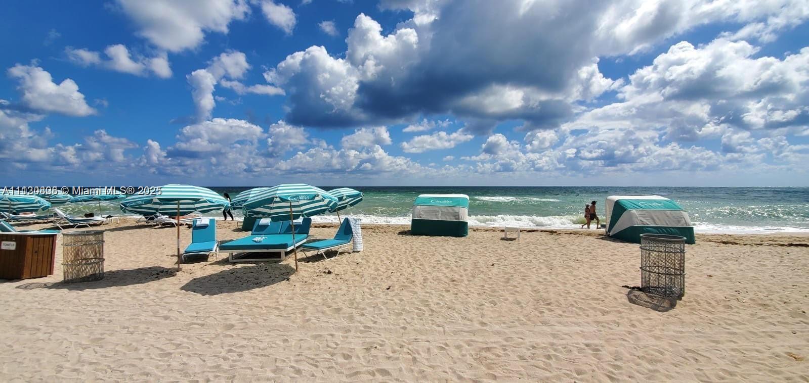 A sandy beach features several striped umbrellas, lounge chairs, and cabanas under a cloudy sky — 6801 Collins Ave # C1115, Miami Beach FL 33141, MIAMI BEACH, FL