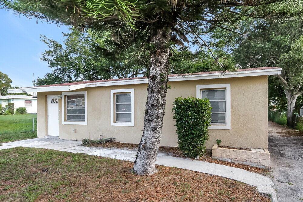 A single-story tan house with a white door and windows sits behind a large tree — RIVIERA BEACH, FL