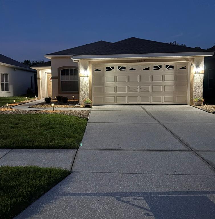 A single-story house with a two-car garage, concrete driveway, and illuminated landscaping at dusk — 23814 CORAL RIDGE LANE, LAND O LAKES, FL