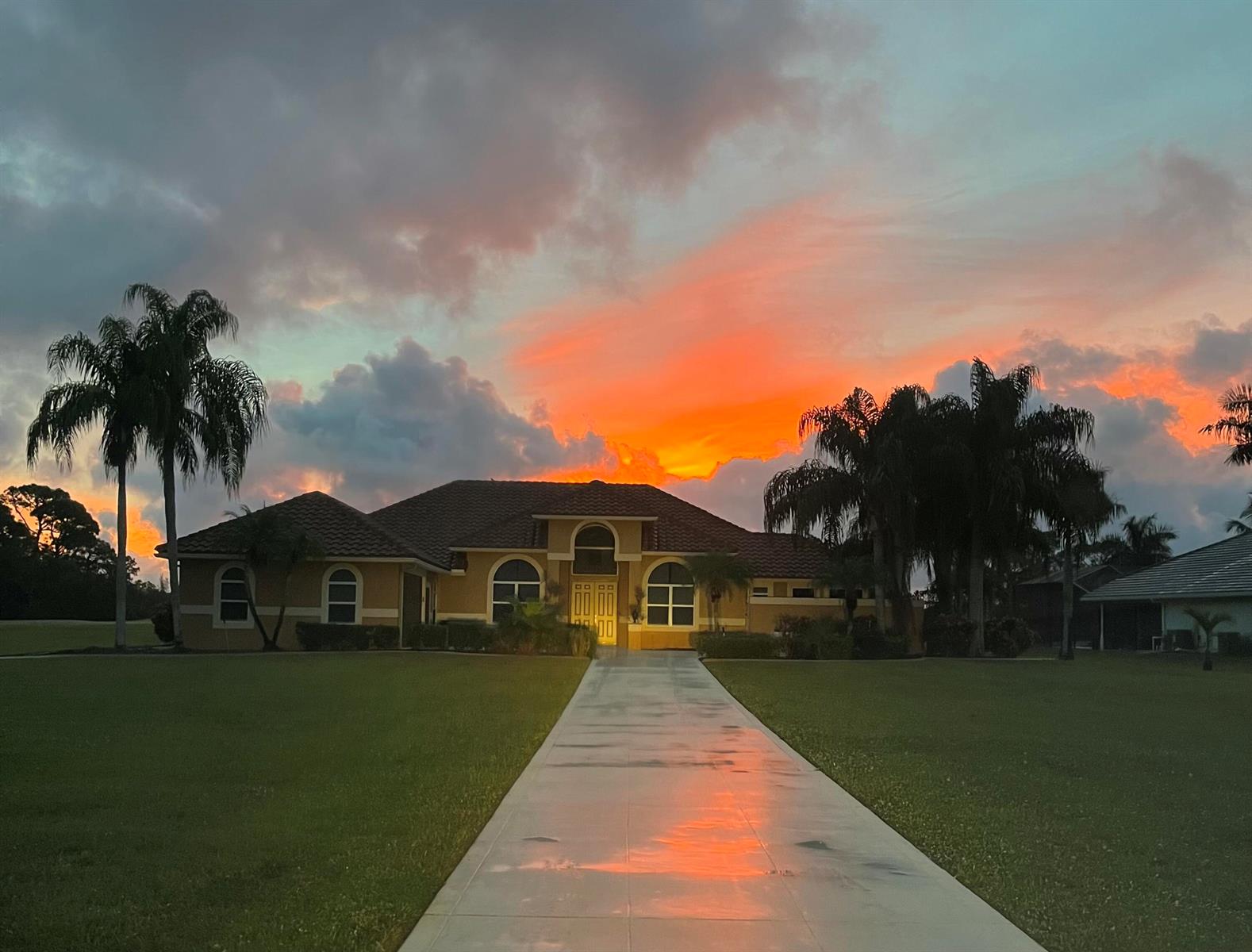 A single-story house with a concrete driveway and palm trees under a vibrant sunset sky — 8030 Woodsmuir Drive, Palm Beach Gardens FL 33412, PALM BEACH GARDENS, FL