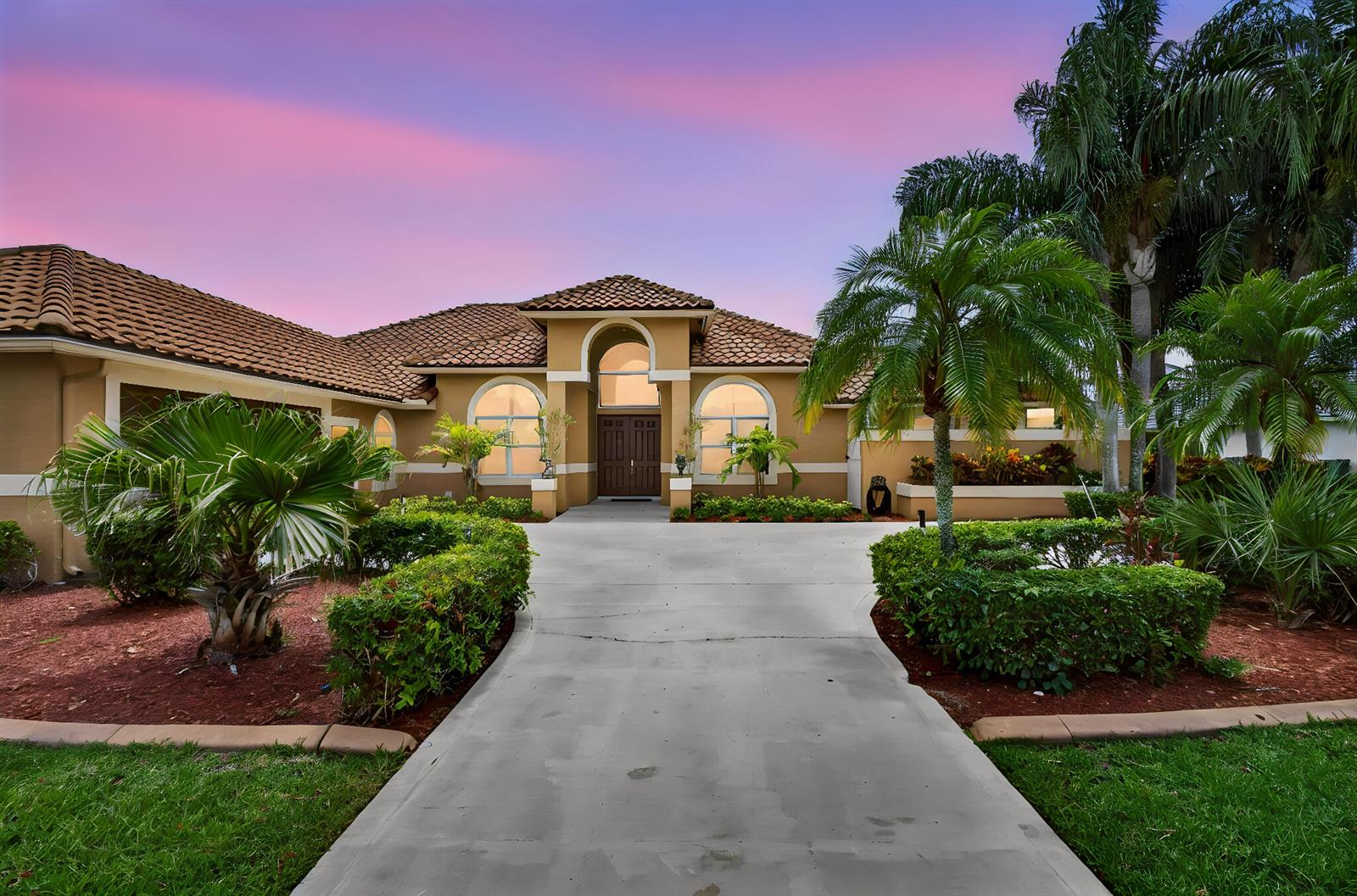 A tan house with a tile roof and arched windows sits behind a concrete driveway — 8030 Woodsmuir Drive, Palm Beach Gardens FL 33412, PALM BEACH GARDENS, FL